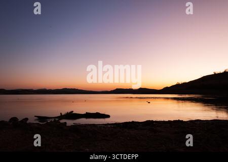 Die untergehende Sonne färbt den Himmel mit leuchtenden Farben und schafft eine friedliche Atmosphäre über dem See. Das Wasser reflektiert den Himmel wie ein Spiegel, was die Ruhe der Szene erhöht Stockfoto