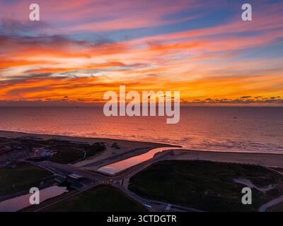 Aus der Vogelperspektive auf den Sonnenuntergang an der Küste mit lebendigem Himmel über dem ruhigen Meeresfluss fließt neben Sandstrand und verstreuten Gebäuden in den Ozean und vermischt warme Li Stockfoto
