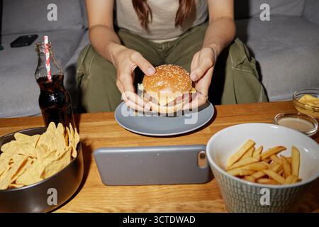 Junge Erwachsene Frau isst Burger, während sie ein Video auf dem Smartphone ansieht Stockfoto