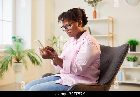 Konzentrierter afroamerikaner-Heim-Sessel, der mit Handy sitzt Stockfoto