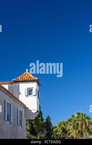 Eine sonnendurchflutete Küstenstraße verfügt über ein historisches Steingebäude mit orangefarbenen Ziegeldächern, Fensterläden und einer von Palmen gesäumten Promenade. Klarer blauer Himmel und s Stockfoto