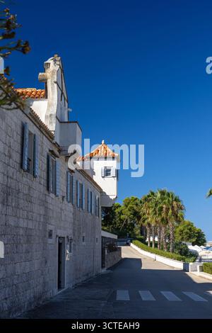 Eine sonnendurchflutete Küstenstraße verfügt über ein historisches Steingebäude mit orangefarbenen Ziegeldächern, Fensterläden und einer von Palmen gesäumten Promenade. Klarer blauer Himmel und s Stockfoto