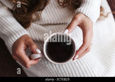 Weibliche Hände halten eine Tasse heißen schwarzen Kaffee in einem weißen Pullover. Gemütliche Morgenstimmung und Entspannung zu Hause. Warmes Getränk und Lifestyle-Komfortkonzept. Stockfoto