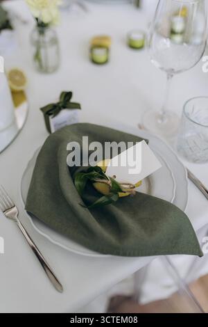 Wunderschön angeordnete Tischdekoration mit grüner Leinenserviette, lebendiger Zitrone, dekorativem Anhänger und charmanten Gefälligkeiten vor einem makellos weißen Ta Stockfoto
