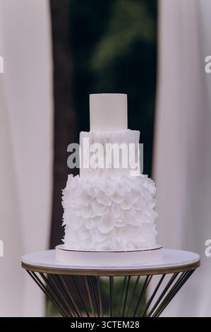 Eine atemberaubende dreistufige weiße Hochzeitstorte, elegant präsentiert auf einem stilvollen Standfuß, verziert mit zarten Rüschen und Blumendetails, perfekt für jeden Geschmack Stockfoto