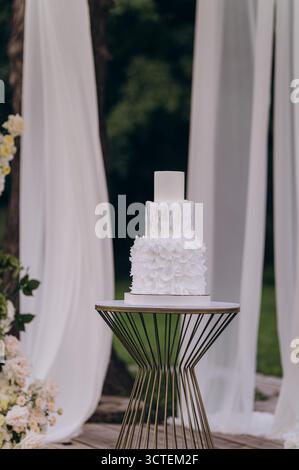 Eine atemberaubende weiße Hochzeitstorte mit zarten floralen Texturen steht prominent auf einem schicken Tisch. Der Hintergrund ist mit weichen Vorhängen und sanften Details versehen Stockfoto