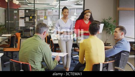 Führende schwarze Frau in blauer Bluse, die Tablette hält, führt Meeting am Tisch im Großraumbüro Stockfoto
