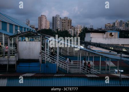 Mumbai, Indien - 23. September 2025: Blick auf eine Fußgängerbrücke über einen Bahnhof, die Skyline der Stadt erhebt sich in der Ferne unter einem dunklen Himmel. Stockfoto
