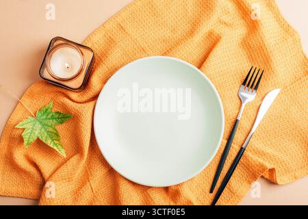 Herbsttisch-Arrangement mit Teller, brennender Kerze, Besteck und gelber Serviette auf beigefarbenem Tisch. Draufsicht, flach, Modell. Stockfoto