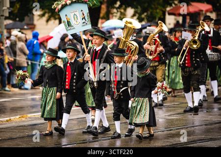München, Deutschland - 18. September 2022: Blick auf Kinder in traditioneller bayerischer Kleidung, die auf dem pulsierenden Oktoberfestzug marschieren, deren bunte Kostüme sich von der nassen Straße abheben. Stockfoto