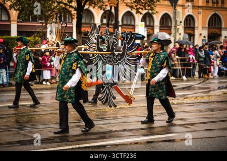 München, Deutschland - 18. September 2022: Blick auf Männer in traditioneller bayerischer Kleidung mit einem großen Adlerwappen durch die Straßen der Stadt, beobachtet von Zuschauern in der Nähe der Frauenkirche. Stockfoto