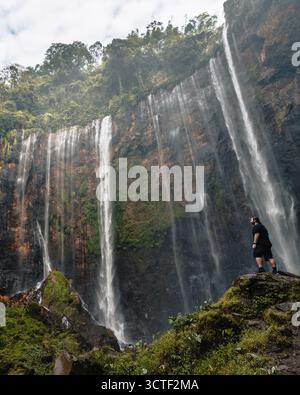 Der Blick auf die majestätischen Tumpak Sewu Wasserfälle stürzt die üppigen grünen Klippen hinunter, eine einsame Figur, die vor der Kraft der Natur steht, Tumpak Sewu, Jawa Tim Stockfoto