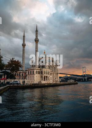 Blick auf die elegante Ortaköy Moschee steht in Harmonie mit dem Bosporus, Brücke Silhouette vor dem farbenfrohen Himmel, reflektiert im Wasser, Istanbul, İstanbul, Türkiye. Stockfoto