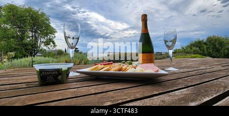 Ein Picknick im Freien mit einer Flasche Champagner, Gläsern und einer Platte mit Snacks auf einem Holztisch mit malerischem Hintergrund. Stockfoto