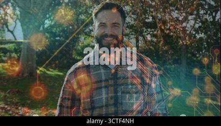 Lächelnder bärtiger Mann in Flanellhemd, der im Garten steht, mit Herbstblättern, glühender Kreisüberlagerung Stockfoto