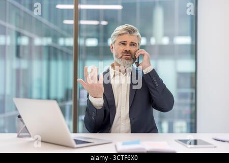 Reifer Geschäftsmann mit grauen Haaren und Bart am Schreibtisch, gestresst und gespenstisch während eines dringenden Telefongesprächs, der Frustration und ernste Bedenken des Unternehmens vermittelt Stockfoto