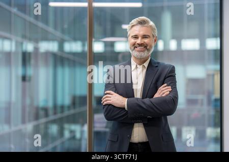 Reifer Geschäftsmann mit grauem Bart und professioneller Kleidung, stehend in einem modernen Büro mit überkreuzten Armen und einem selbstbewussten, freundlichen Lächeln Stockfoto