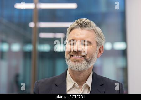 Ausdrucksstarker Geschäftsmann mit grauem Haar und Bart, der die Augen schließt und lächelt, spiegelt Freude, Glück und Erfolg in einer modernen Büroumgebung wider Stockfoto