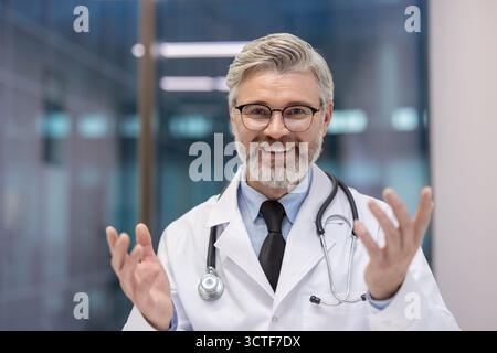Leitender männlicher Arzt mit Brille und weißem Mantel lächelnd, redend, und Gesten, arbeitet in einem Krankenhaus oder einer Klinik, repräsentiert Gesundheitswesen, Medizin und professionelle Expertise Stockfoto