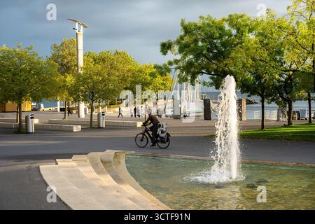 Lausanne, Schweiz - 24. September 2025: Ein Radfahrer fährt an einem Brunnen vorbei in einem sonnigen Park von Lausanne Ouchy mit Bäumen und Blick aufs Wasser. Stockfoto