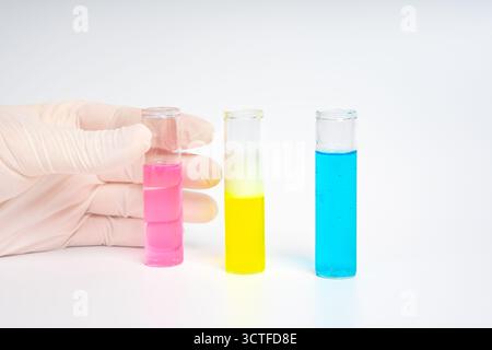 Hände mit Handschuhen, die Glasröhrchen mit blauen, rosa und gelben Flüssigkeiten auf isoliertem weißen Hintergrund halten. Farbenfrohe wissenschaftliche Experimente. Chemische Reaktion Stockfoto