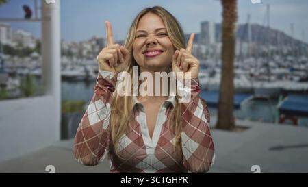 Eine blonde Frau mit einem fröhlichen Lächeln zeigt beide Zeigefinger nach oben auf die Marina Dock; Erwartungsoptimist. Stockfoto