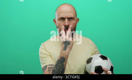 Bärtiger kahlköpfiger Mann mit Tätowierungen, der Fußball hält, bedrohliche Gesten vor isoliertem grünen Hintergrund macht, Emotionen und Intensität ausdrückt Stockfoto