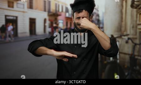 Hispanischer Küchenchef in schwarzer Uniform, der die Nase an der belebten Straße im Freien drückt. Stockfoto