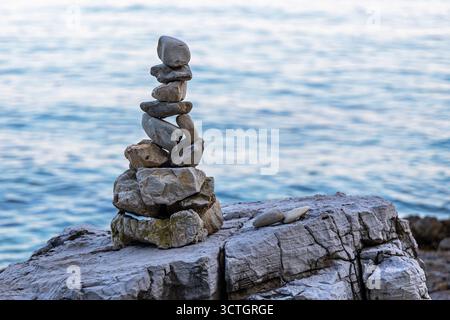 Ausgeglichener Steinstapel am Meer. Sorgfältig gestapelte Steine bilden einen ausgeglichenen Turm an einem felsigen Ufer mit ruhigem blauem Meerwasser im Hintergrund. Stockfoto