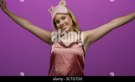 Frau lächelt in pinkfarbener Nachtwäsche und Katzenohrmaske vor leuchtendem lila Hintergrund, drückt Freude und Entspannung aus und hält positive und verspielte ener fest Stockfoto