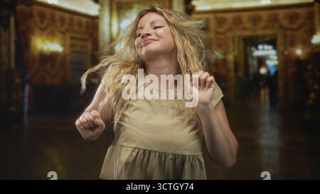 Frau tanzt mit geschlossenen Augen und erhobenen Händen in einem kunstvollen Museumssaal, der mit Kronleuchtern und vergoldeten Säulen gesäumt ist; Glücksbewegung. Stockfoto