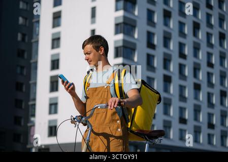 Junger Mann, der als Fahrradkurier arbeitet und sein Smartphone auf Wegbeschreibungen oder Bestellungen überprüft, während er mit seinem Fahrrad steht, Lebensmittel oder Pakete in der Stadt liefert Stockfoto