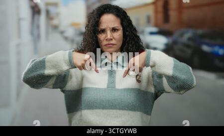 Frau mit lockigem Haar und Brille, die auf einer städtischen Straße steht und einen gestreiften Pullover trägt, zeigt selbstbewusst auf sich selbst in diesem Outdoor-Settin Stockfoto