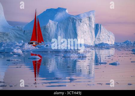 Ein markantes rotes Segelboot gleitet durch ruhige arktische Gewässer, umgeben von riesigen Eisbergen bei Sonnenuntergang in Grönland. Das warme Abendlicht reflektiert das Licht Stockfoto