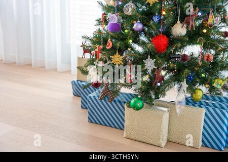 Geschenkverpackungen unter einem geschmückten Weihnachtsbaum mit leuchtenden Ornamenten und festlichen Lichtern zu Hause. Stockfoto