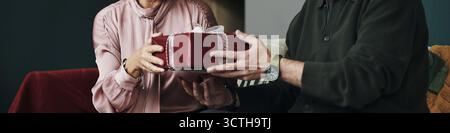 Kopfzeile einer älteren Frau, die eine Geschenkbox vom Ehemann erhält, beide sitzen dicht beieinander auf dem Sofa, Hände berühren, während sie Geschenke austauschen, Gesichter teilweise sichtbar Stockfoto