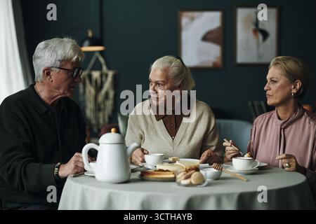Drei ältere kaukasische Leute sitzen am runden Tisch und essen Dessert und trinken Kaffee, zwei Frauen und ein Mann, die sich unterhalten, Gebäck und Teekanne auf dem Tisch während des freundlichen Treffens Stockfoto