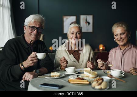 Drei ältere kaukasische Leute sitzen am Tisch und genießen Dessert, zusammen Tee, lächeln und in die Kamera schauen, Tassen und Teller mit Gebäck und Brot halten und einen freundlichen Moment teilen Stockfoto