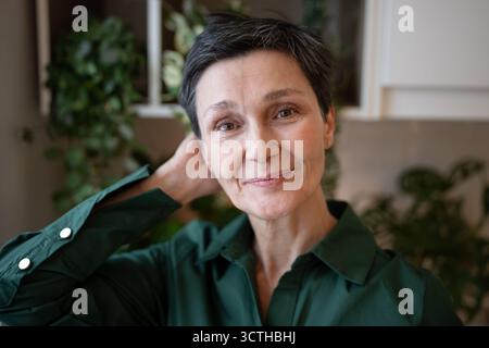 Lächelnde reife Frau, die in die Kamera schaut, glättet die Haare. Selbstbewusstes weibliches Porträt, glücklicher Senior Stockfoto