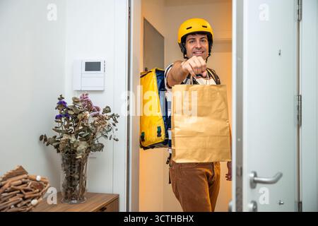 Ein lächelnder Liefermann in gelbem Helm und Thermorucksack überreicht eine Lebensmittelbestellung aus braunem Papier vor einer Haustür, die schnelle, bequeme HO bietet Stockfoto