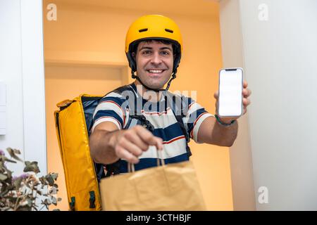 Lächelnder hispanischer Liefermann mit gelbem Helm und großem gelben Thermorucksack, mit einer Papiertüte zum Mitnehmen und einem Stockfoto