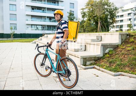 Hispanischer Mann, der einen Schutzhelm und einen gelben Rucksack trägt, neben seinem Fahrrad steht und aktiv als Kurier in einem Modus arbeitet Stockfoto