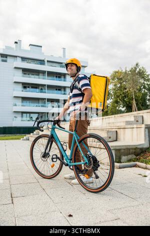 Hispanischer Mann in einem gestreiften T-Shirt und Sicherheitshelm, der mit seinem modernen Fahrrad und der gelben Liefertasche steht und Dienstleistungen in einer städtischen Umgebung erbringt Stockfoto