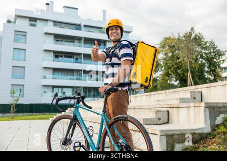 Glücklicher spanischer Liefermann, der einen gelben Helm trägt und einen Lieferrucksack trägt, mit seinem Fahrrad steht und in einer Stadt ein Daumen nach oben zeigt Stockfoto