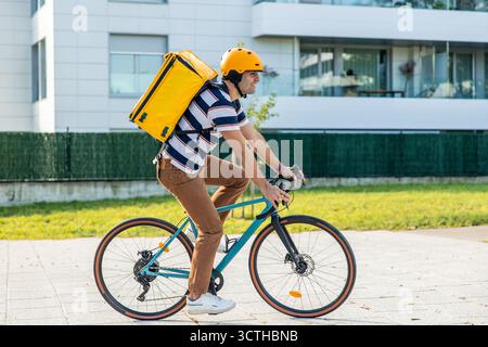 Hispanischer Liefermann mit gelbem Helm und großem, gelbem Thermalrucksack, Fahrrad auf einem Bürgersteig in urbaner Umgebung, Bringi Stockfoto