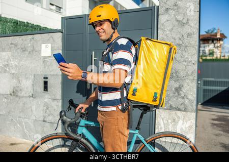 Ein spanischer Liefermann, der einen Helm trägt und einen gelben Thermorucksack trägt, überprüft sein Smartphone neben einem Fahrrad in einer Stadt und liefert Lebensmittel an einen Stockfoto
