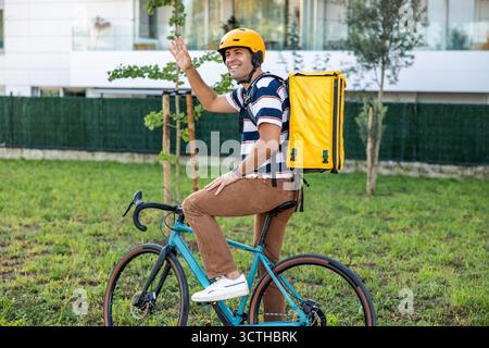 Hispanische Lieferarbeiter im Helm, lächelnd und winkend, während sie auf dem Fahrrad mit gelber Thermotasche sitzen, repräsentiert einen schnellen, umweltfreundlichen Kurier in der Stadt Stockfoto