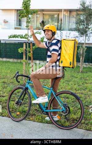 Hispanischer Zustellkurier in gelbem Helm und Rucksack lächelt und weht, während er auf einem Fahrrad auf einer sonnigen Wohnstraße sitzt, umweltfreundlich, städtisch Stockfoto