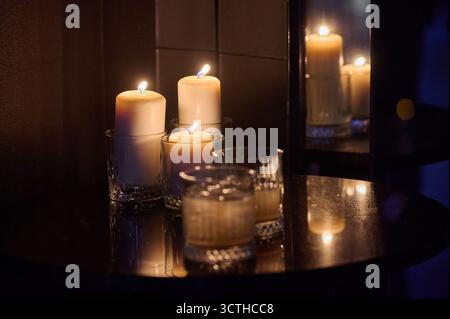 Drei beleuchtete Kerzen in Glashaltern auf reflektierendem Tisch stimmungsvolle Ambiente Inneneinrichtung kommerzielle Produktaufnahme Stockfoto