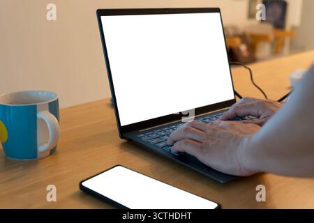 Nahaufnahme der Hände, die auf einem Laptop mit leerem weißen Bildschirm neben dem Smartphone und einer Kaffeetasse auf einem Holztisch tippen. Modernes Arbeitsplatztechnologiekonzept für d Stockfoto